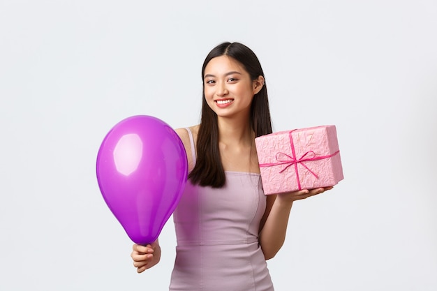 Celebrazione, festa e concetto di vacanze. Bella ragazza asiatica in abito da sera, sorridendo compiaciuto, godendo di festeggiare il compleanno, tenendo in mano un regalo di compleanno e un palloncino