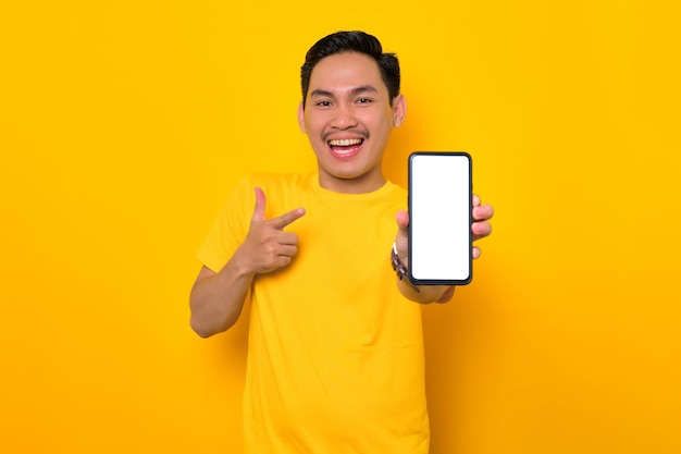 Cellulare mockup schermo vuoto Giovane asiatico allegro in maglietta casual che mostra il telefono cellulare con schermo vuoto che consiglia app mobile isolata su sfondo giallo Concetto di stile di vita delle persone