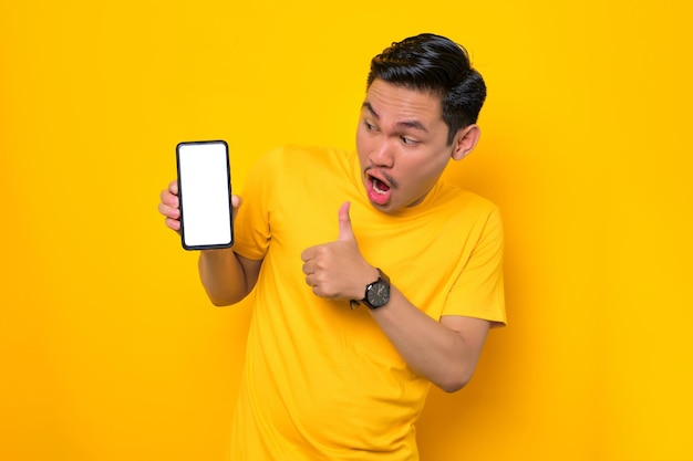 Cellulare mockup schermo vuoto Giovane asiatico sorpreso in maglietta casual che mostra il telefono cellulare con schermo vuoto che mostra il pollice in alto gesto isolato su sfondo giallo Concetto di stile di vita delle persone