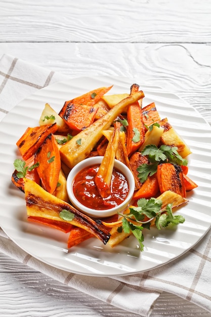 Cena arrosto domenicale: pastinaca e carota arrosto con spezie ras el hanout: cannella, curcuma, anice su un piatto bianco con ketchup, coriandolo fresco su un tavolo di legno bianco, vista dall'alto, primo piano