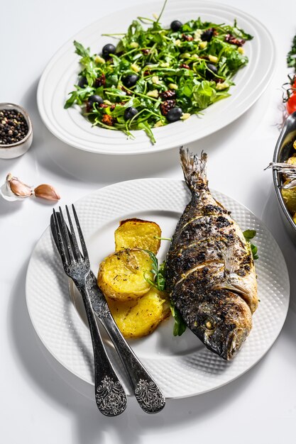 Cena fatta in casa con pesce Dorado al forno, insalata di rucola con pomodori, patate al forno. Sfondo bianco. vista dall'alto