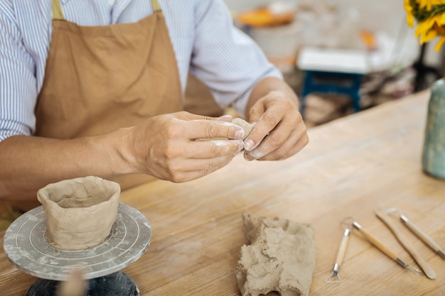 Ceramista e oggetti. Ceramista che indossa un grembiule marrone seduto al tavolo da lavoro in legno che forma piccoli oggetti