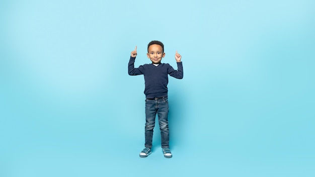 Cerca Adorabile bambino afroamericano che punta le dita verso l'alto nello spazio libero e sorride alla fotocamera a tutta lunghezza