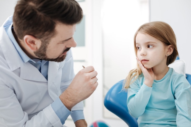 Cerchiamo di lavorare in squadra. Piacevole dentista professionista pediatrico che dice alla bambina che ha bisogno del suo aiuto per capire la causa del dolore ai denti