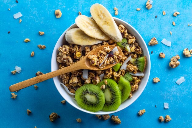Cereali per la colazione a secco. Ciotola croccante di muesli del miele con le fette di banana e di kiwi freschi su un fondo blu. Cibo sano, fitness e fibra. Vista dall'alto. Ora di colazione