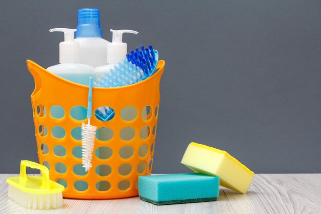 Cestino in plastica con bottiglie di detersivo per piatti, detergente per vetri e piastrelle, detergente per forni a microonde e fornelli con spazzola e spugne su sfondo grigio. Kit di lavaggio e pulizia.