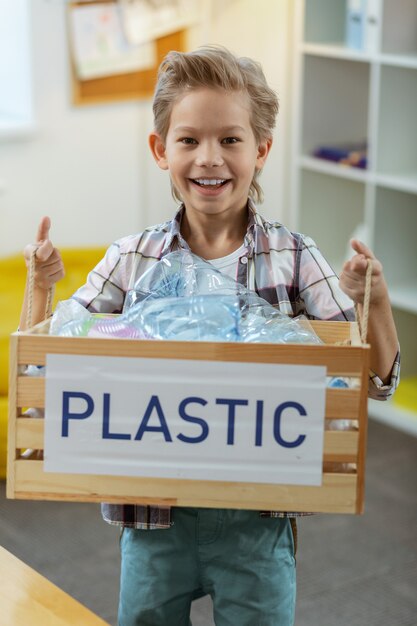 Cestino per il riciclaggio. Ragazzino sorridente in camicia a scacchi che presenta una scatola con bottiglie di plastica come risultato di un duro processo di separazione