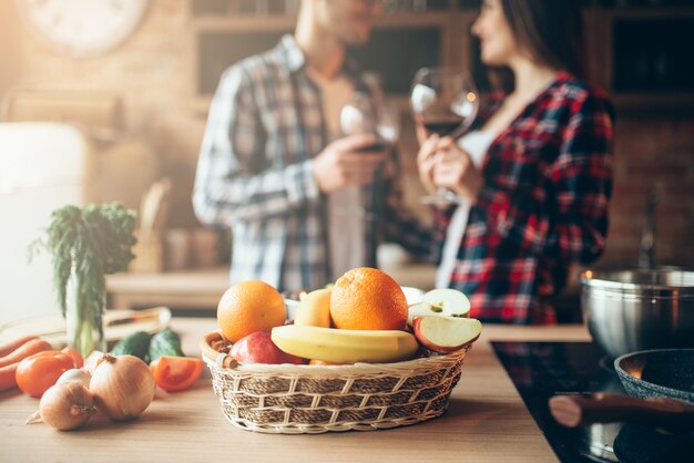 Cesto di frutta fresca sul tavolo, coppia di innamorati che beve vino in cucina a casa, cena romantica in famiglia