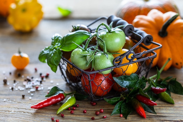 Cesto di metallo con diversi pomodori rossi, gialli e verdi maturi su un tavolo di legno e foglie di basilico