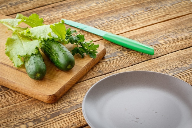 Cetrioli appena raccolti e insalata verde su tagliere, coltello e piatto su vecchie tavole di legno. Verdure appena raccolte. Vista dall'alto.