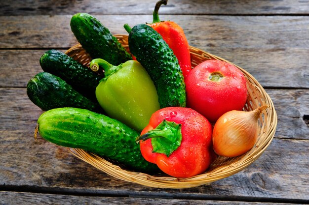 Cetrioli, pomodori, peperoni dolci e cipolle in un cesto su un tavolo di legno in stile rustico.