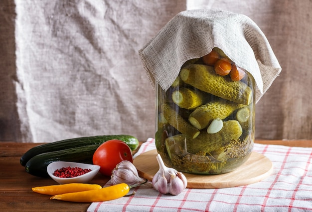 Cetrioli sottaceto e pomodori in un vaso di vetro sulla tovaglia di lino e tavolo in legno.