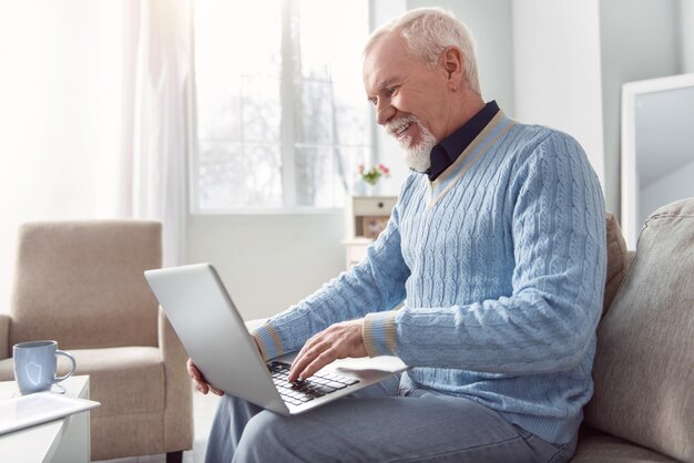 Chat che stimola l'umore. Allegro uomo anziano seduto sul divano e digitando un messaggio di posta elettronica sul laptop mentre sorride felicemente