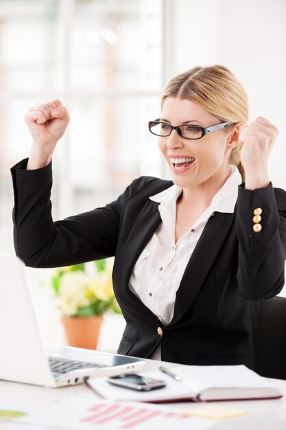 Che giorno fortunato! Felice imprenditrice matura seduta al suo posto di lavoro e gesticolando