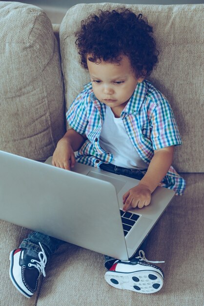 Che pulsante devo premere? Veduta dall'alto del piccolo bambino africano che usa il suo laptop mentre è seduto sul divano di casa