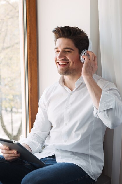 che ride felice uomo che indossa una camicia bianca utilizzando computer tablet e cuffie mentre era seduto alla finestra nel soggiorno