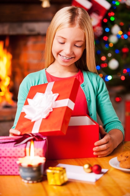 Che sorpresa! Bambina carina che apre una scatola regalo e sorride mentre è seduta al tavolo con l'albero di Natale e il caminetto sullo sfondo