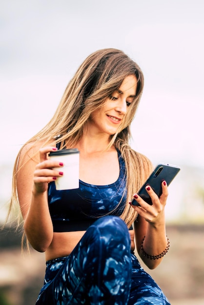 Cheerful e bella giovane donna caucasica sana pausa dopo l'allenamento di fitness all'aperto e bere un caffè mentre guardare e utilizzare moderno dispositivo di telefono mart persone e concetto di stile di vita di salute