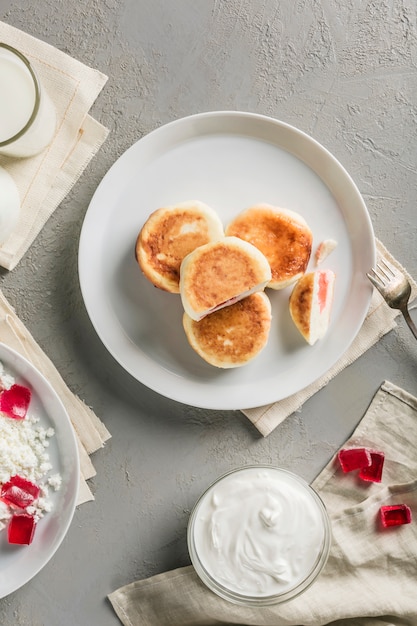 Cheesecakes di ricotta con marmellata di lamponi su un piatto con panna acida e un bicchiere di latte.