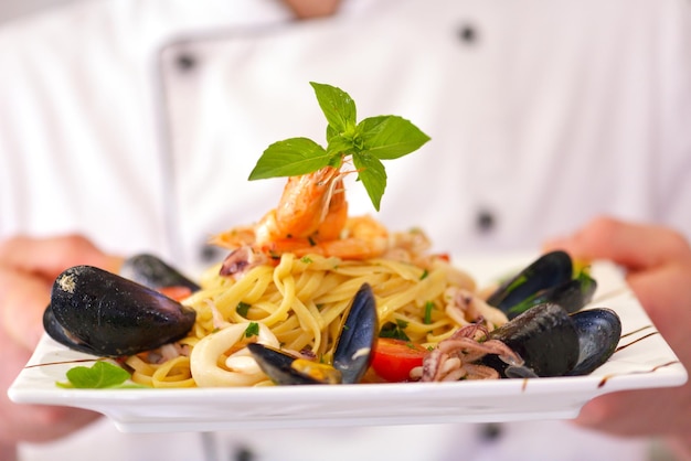 Chef bello vestito in uniforme bianca che decora insalata di pasta e pesce di mare in una cucina moderna