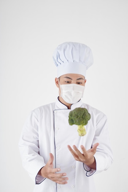 Chef del ristorante in maschera medica che lancia broccoli isolati su bianco