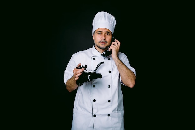 Chef di cucina che indossa giacca e cappello da cucina, parla su un telefono retrò, su sfondo nero