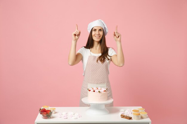 Chef donna casalinga cuoco pasticcere o fornaio in t-shirt bianca grembiule, cappello da chef toque che cucina torta o cupcake a tavola isolato su sfondo rosa pastello in studio. Mock up copia spazio concetto di cibo.