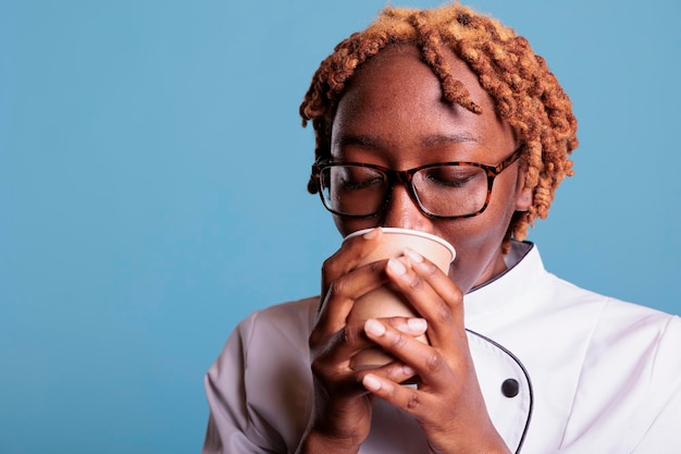 Chef femminile di successo che beve caffè indossando l'uniforme da cucina mentre si prende una pausa da una lunga giornata di lavoro. Membro del personale di cucina femminile in studio girato su sfondo blu.