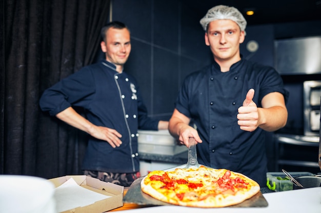 Chef fornaio cuoco in uniforme nera che mette la pizza nel forno con la pala nella cucina del ristorante.