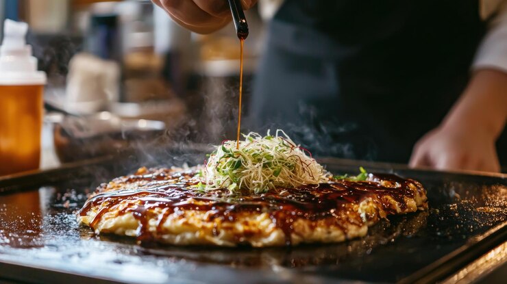 Chef grilling okonomiyaki on a hotplate drizzling sauce on top