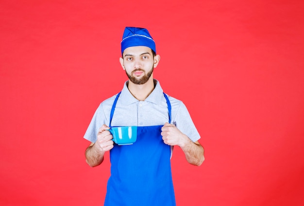 Chef in grembiule blu che tiene una tazza di ceramica blu e mostra il pugno.