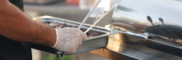 Chef in guanti di gomma che tengono vassoio di metallo con cibo e pinze in primo piano