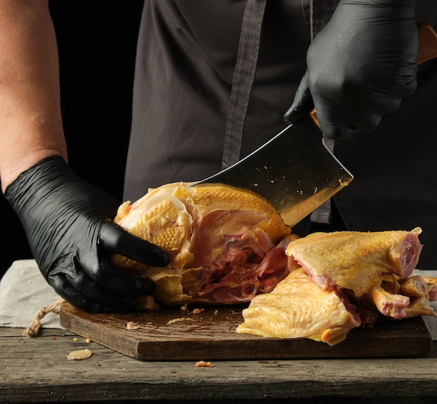 Chef in guanti di lattice e uniforme nera che tagliano a pezzi il pollo crudo su una tavola di legno marrone