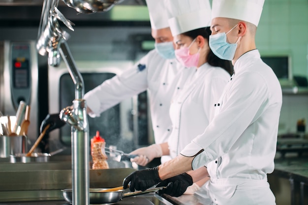 Chef in guanti e maschere protettive preparano il cibo nella cucina di un ristorante o di un hotel.