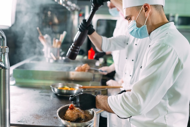 Chef in guanti e maschere protettive preparano il cibo nella cucina di un ristorante o di un hotel.