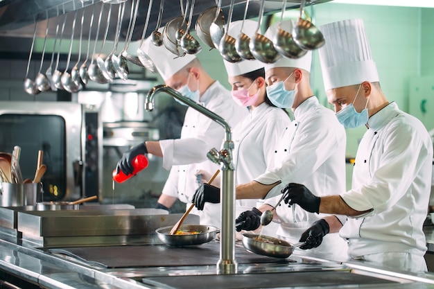 Chef in guanti e maschere protettive preparano il cibo nella cucina di un ristorante o di un hotel.