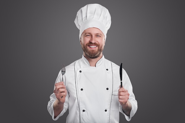 Chef maschio barbuto adulto positivo in uniforme bianca che tiene forchetta e coltello e che guarda l'obbiettivo mentre rappresenta il servizio di ristorante su sfondo grigio