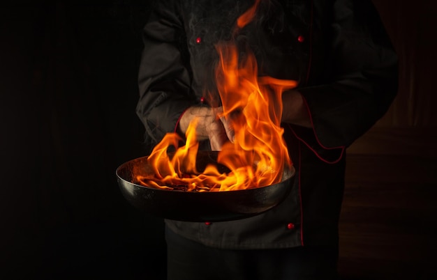 Chef professionista che cucina cibo in padella con fiamma di fuoco su sfondo nero