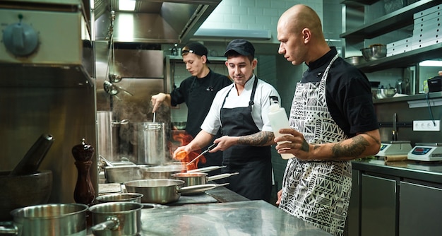 chef professionista che insegna ai suoi due giovani apprendisti come flambare il cibo in modo sicuro. tecniche di cottura