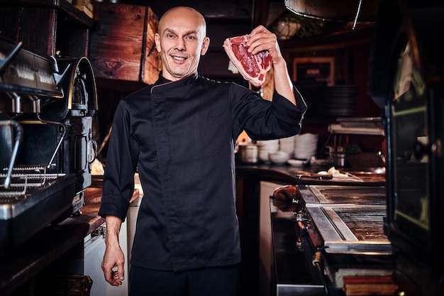 Chef sorridente che indossa l'uniforme con in mano un pezzo di carne fresca e guardando una telecamera nella cucina di un ristorante.