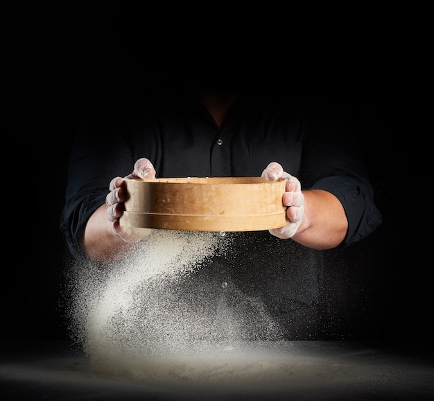 Chef un uomo in uniforme nera tiene un setaccio di legno rotondo