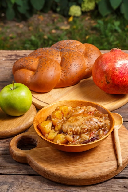 Chelnt con pollo in un piatto di legno con un cucchiaio di legno sul tavolo festivo per Rosh Hashanah accanto alle mele challah e melograno
