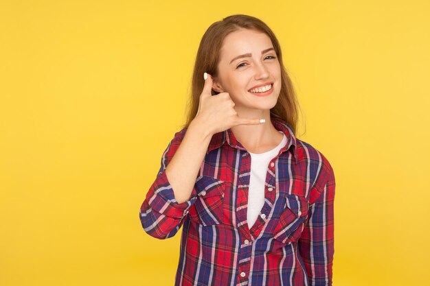 Chiamami Ritratto di dolce adorabile bella ragazza in camicia a scacchi che fa gesto telefonico vicino all'orecchio e sorridente in attesa di contatto di chiamata per telefono studio indoor girato isolato su sfondo giallo