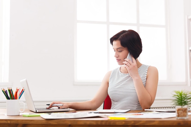 Chiamata importante. Giovane donna di affari seria nel parlare casualmente sul telefono cellulare nell'interno bianco moderno dell'ufficio, spazio della copia