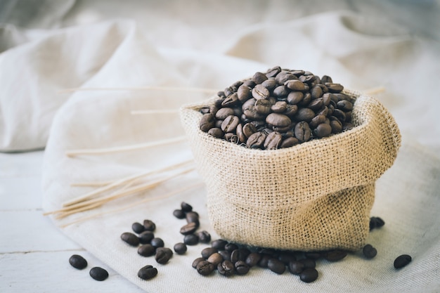 Chicchi di caffè in sacco di iuta e bastone su fondo di legno bianco