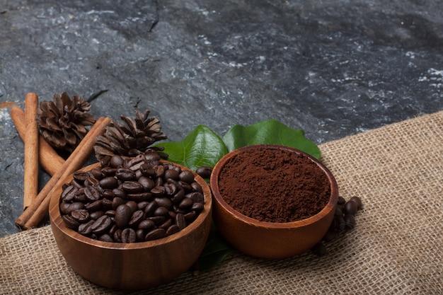 Chicchi di caffè in tazza di legno sulla foglia verde, pino su tela da imballaggio su pietra nera strutturata