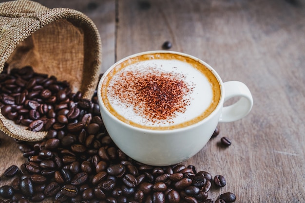 Chicchi di caffè nel sacco con una tazza di caffè sul fondo della tavola in legno.