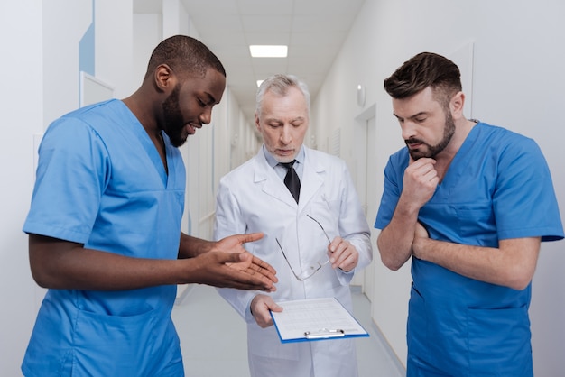 Chiedere una consulenza professionale. Stagista afroamericano ispirato positivo in piedi in ospedale mentre discute la diagnosi con colleghi esperti e si gode la conversazione