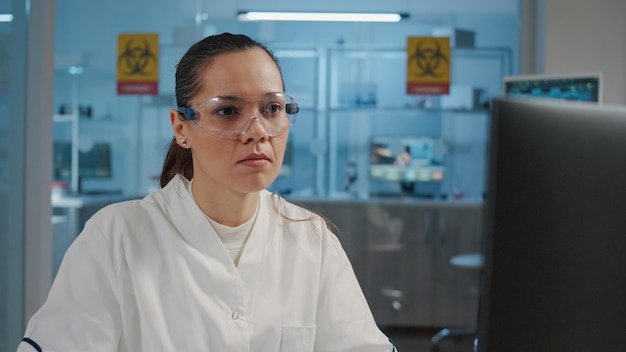 Chimico donna che lavora al computer nel laboratorio di scienze, utilizzando la tecnologia per sviluppare esperimenti di microbiologia con apparecchiature di laboratorio. Scienziato con occhiali protettivi. Scatto a mano libera