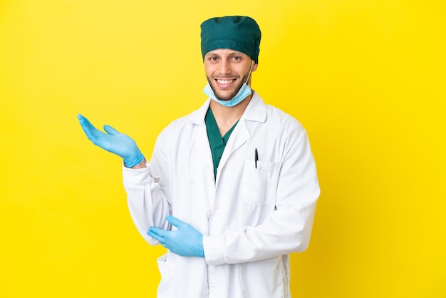 Chirurgo biondo in uniforme verde isolato su sfondo giallo che estende le mani di lato per invitare a venire